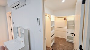 Habitación de hotel con tocador blanco, espejo, aire acondicionado integrado, panel de control y armario con estantes.