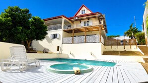 Un edificio a due piani con piscina, terrazza in legno e zona salotto all'aperto coperta.