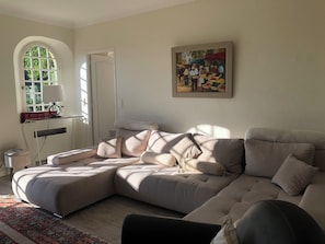 A living room with a sectional sofa, a painting, a window with books, and a lamp.