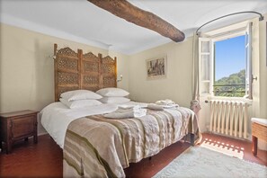A bedroom with a bed, a wooden headboard, a window with a view, a radiator, and a small table. - Mas provençal with swimming pool on 3 hectares of terraced land. Pets welcome (Cabris)