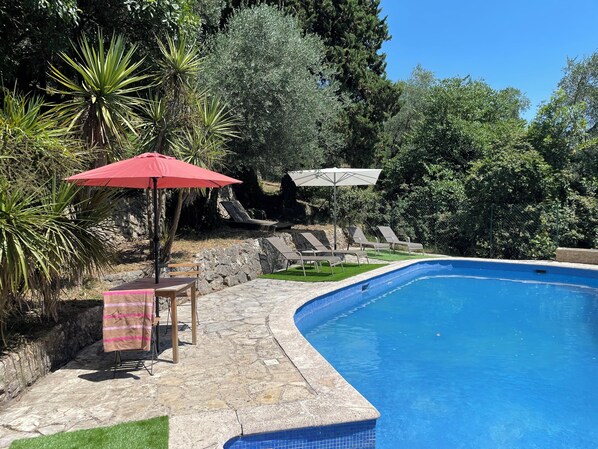 A swimming pool with a stone deck, lounge chairs, and an umbrella.