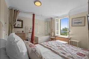A bedroom with a bed, a window with a view, a bathroom with a bathtub, and a chair.