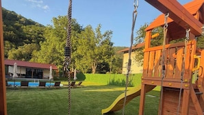 A wooden swing set with red seats and a slide, set against a backdrop of a grassy yard and a building with a red roof.