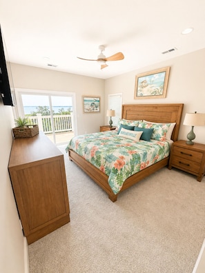 A bedroom with a bed, a ceiling fan, a nightstand, a lamp, a wooden desk, and a view of the outdoors.