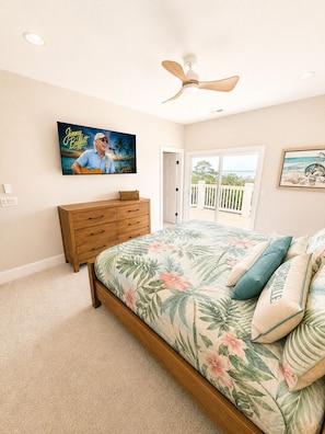 A bedroom with a bed, a ceiling fan, a TV on the wall, a wooden dresser, and a view through a door.