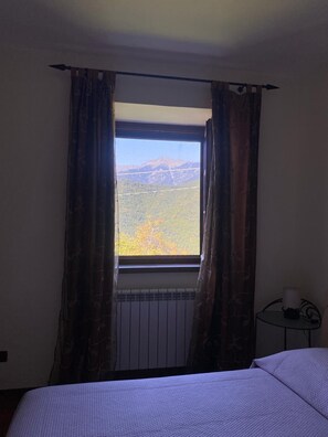 A room with a bed, a window with curtains, a radiator, and a small table with a cup.
