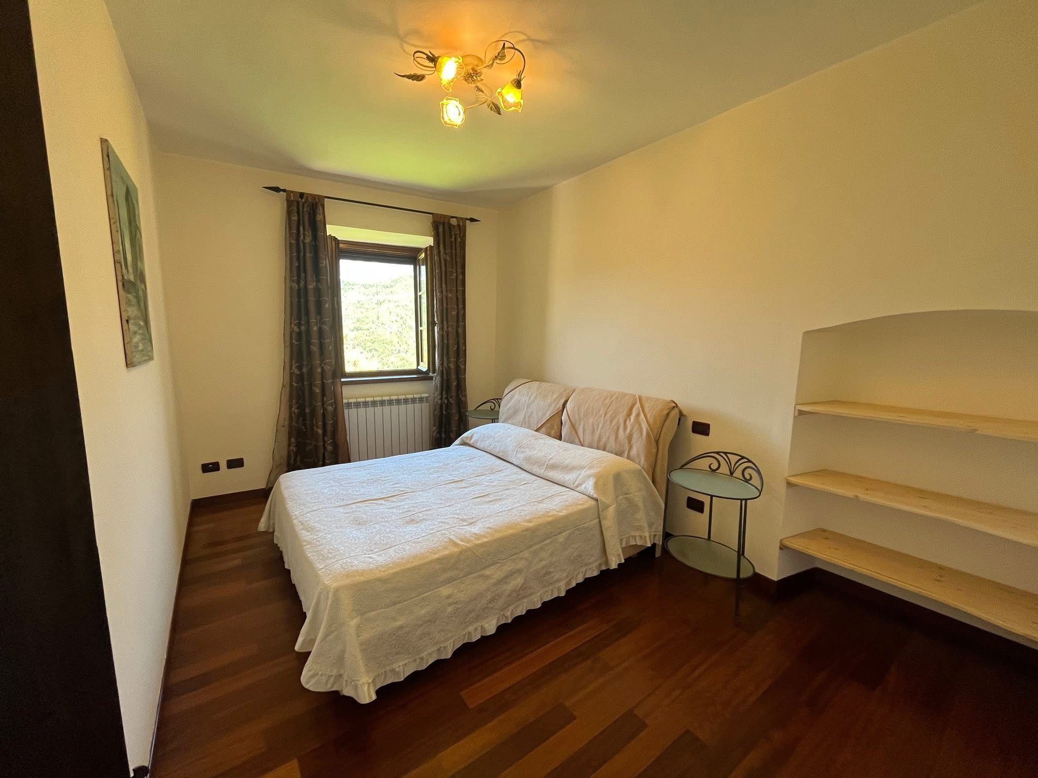 Una camera da letto con un letto, una finestra con tende, un termosifone, un tavolino e una scaffalatura.
