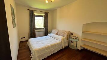 Una camera da letto con un letto, una finestra con tende, un termosifone, un tavolino e una scaffalatura.