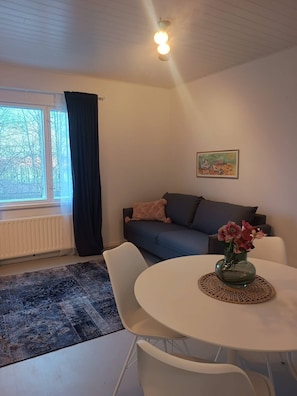 A living room with a sofa, a dining table with chairs, a radiator, a window with curtains, and a painting on the wall. - Cozy Apartment Near the Center (Hameenlinna)