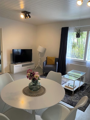 A modern living room with a flat-screen TV, a round dining table with chairs, a glass coffee table, a floor lamp, and a view of greenery through a window.