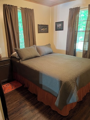A bedroom with a bed, curtains, a framed picture, and a wooden floor.
