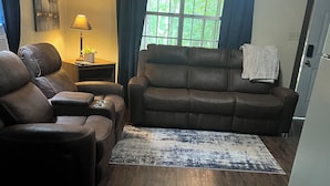A living room with a brown leather sofa, a smaller armchair, a rug, and a window with curtains.