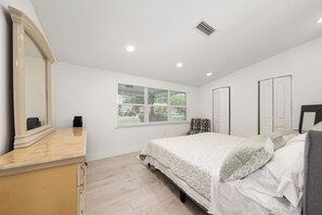 Een slaapkamer met een bed, een dressoir, een raam met jaloezieën en een spiegel.