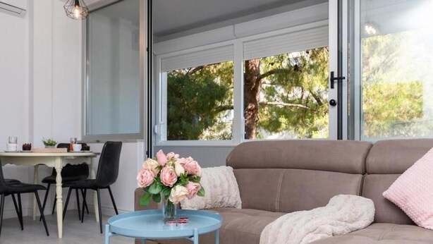 A modern living room with a sofa, a small round table with a vase of flowers, a dining table with chairs, and large windows with a view of trees. - Leora Deluxe Apartment  (Split)