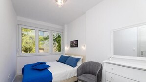 A bedroom with a bed, a chair, a dresser, and a window with a view of greenery. - Leora Deluxe Apartment  (Split)