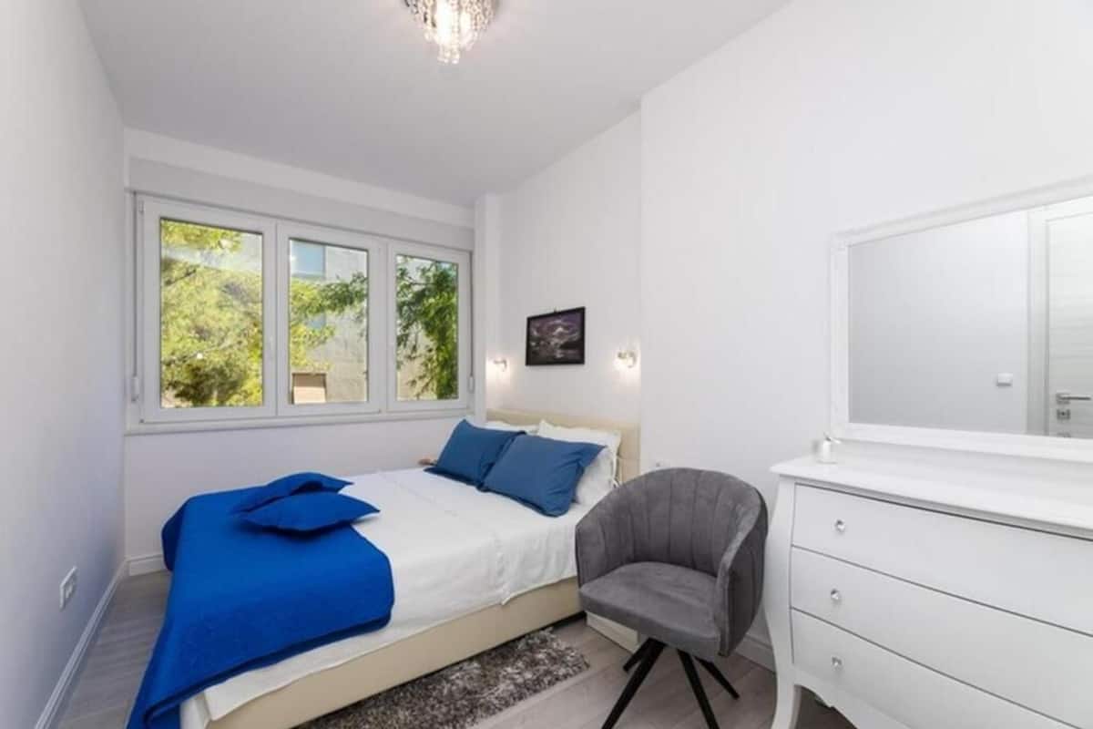A bedroom with a bed, a chair, a dresser, and a window with a view of greenery.