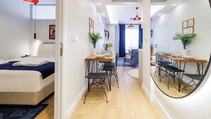 Habitació d'hotel moderna amb un llit, una zona de menjador amb taula i cadires, un mirall gran i un passadís que condueix a una sala d'estar.