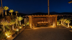 Un espace de restauration en plein air agrémenté d’une pergola, de tables, de chaises et offrant une vue sur le paysage urbain illuminé la nuit.