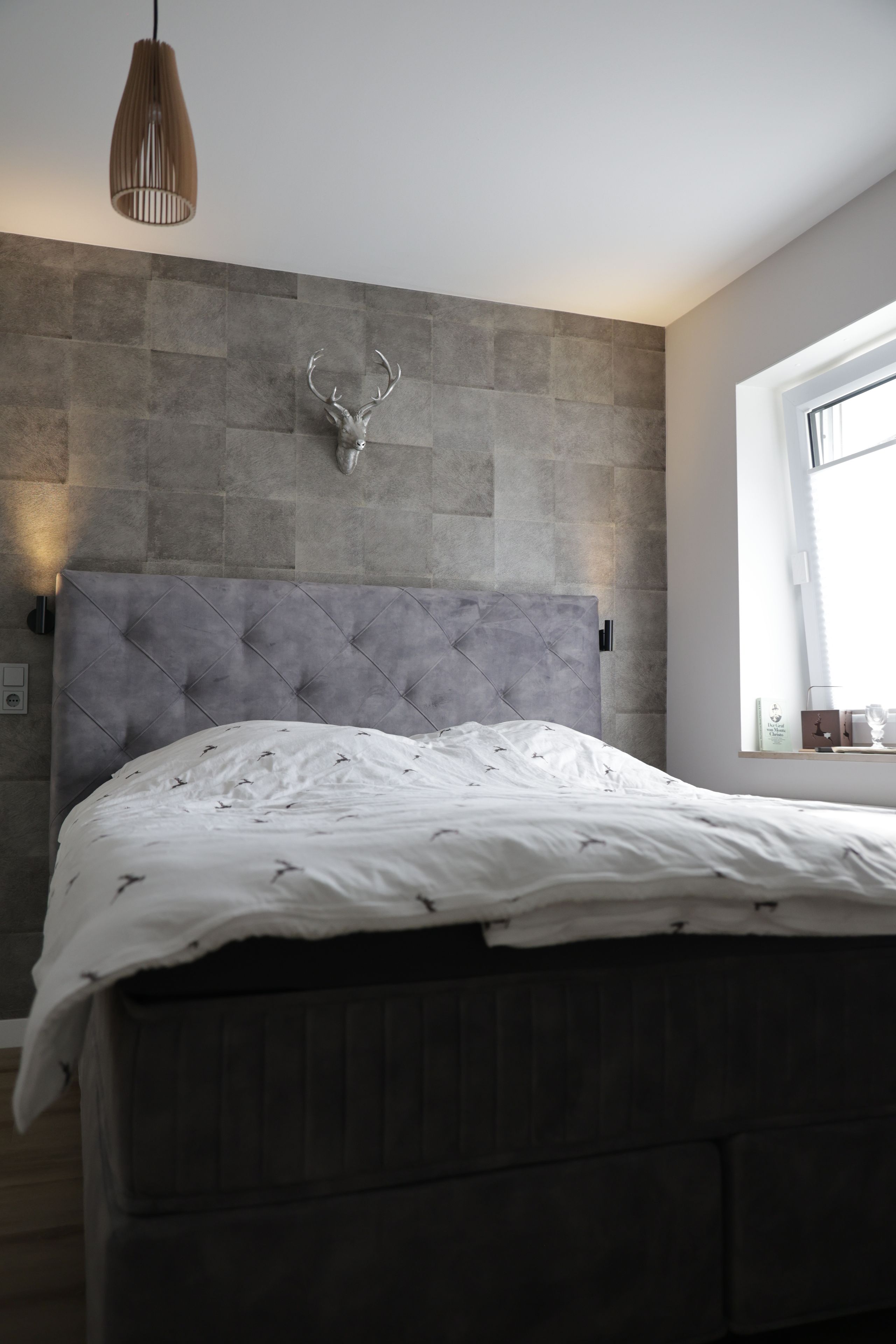 Ein Schlafzimmer mit grau gepolstertem Kopfteil, einem aufgehängten Hirschgeweih an der Wand und einem Fenster mit Jalousien.