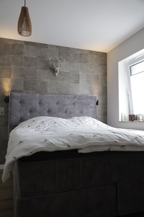 Ein Schlafzimmer mit grau gepolstertem Kopfteil, einem aufgehängten Hirschgeweih an der Wand und einem Fenster mit Jalousien.