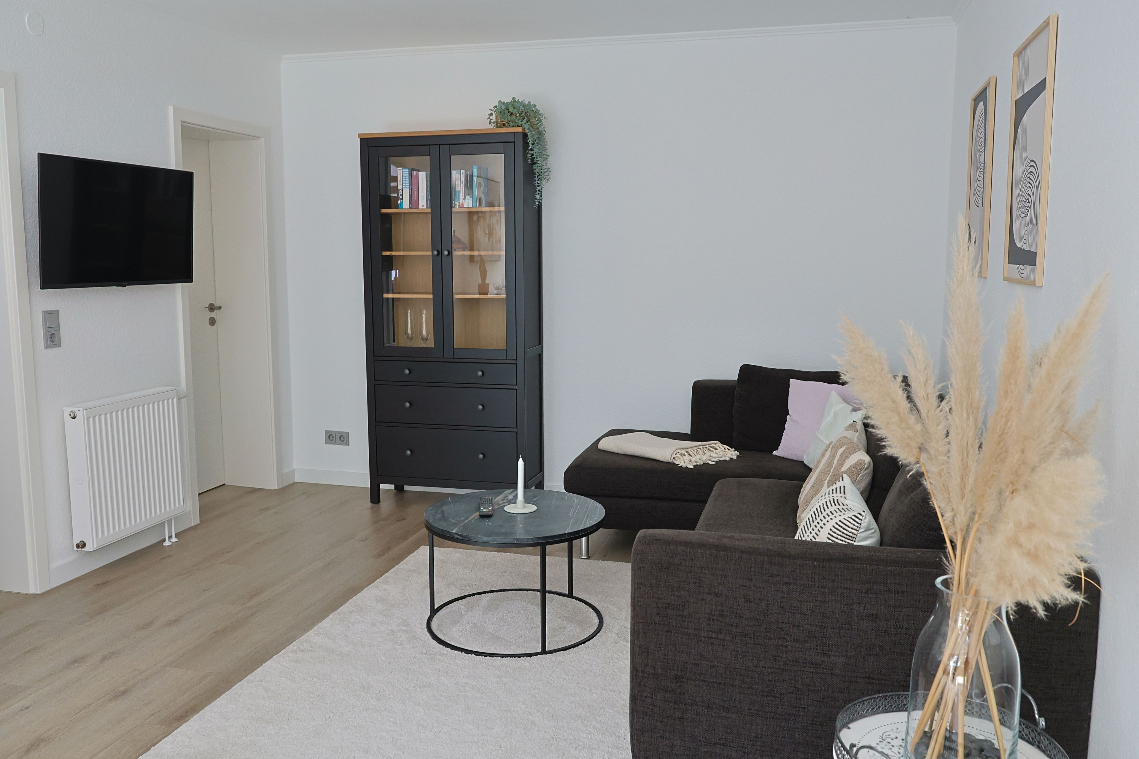 Ein Wohnzimmer mit einem dunkelgrauen Sofa, einem runden Couchtisch, einem an der Wand befestigten Fernseher, einer Vitrine mit Glastüren und einem Heizkörper am Boden.