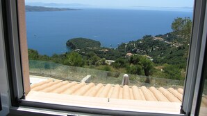 A view from a window overlooking a coastal landscape with a clear sky, a body of water, and a hilly terrain with scattered buildings. - Stone Villa Vasiliki Private Pool & Sea View (Corfu)
