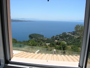 Blick aus einem Fenster auf eine Küstenlandschaft mit blauem Himmel, einem Gewässer und hügeligem Gelände mit verstreuten Gebäuden.