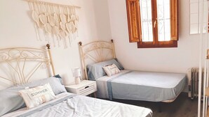 Habitació amb dos llits, una taula de nit amb una làmpada, un penjoll macramè i una finestra amb persianes de fusta.