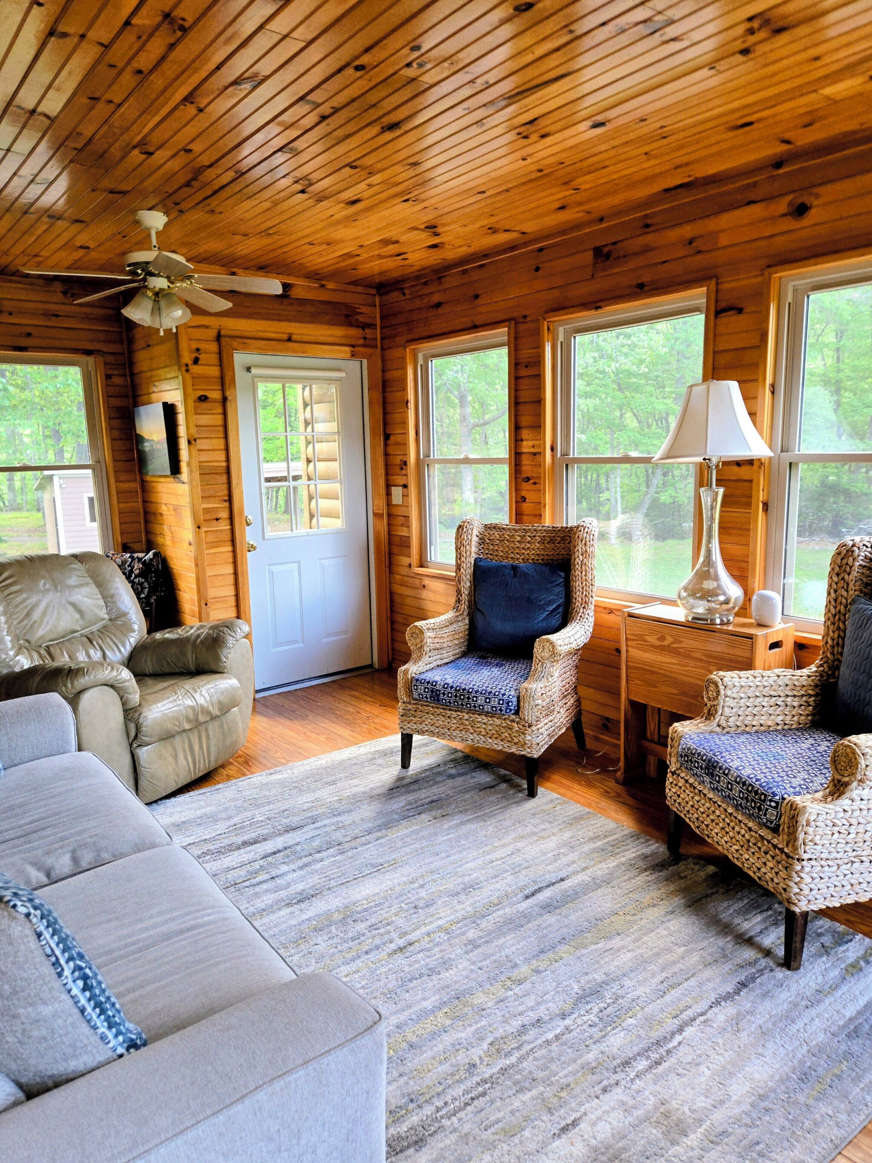 A wooden cabin interior with a ceiling fan, two wicker chairs, a sofa, and a desk with a lamp.