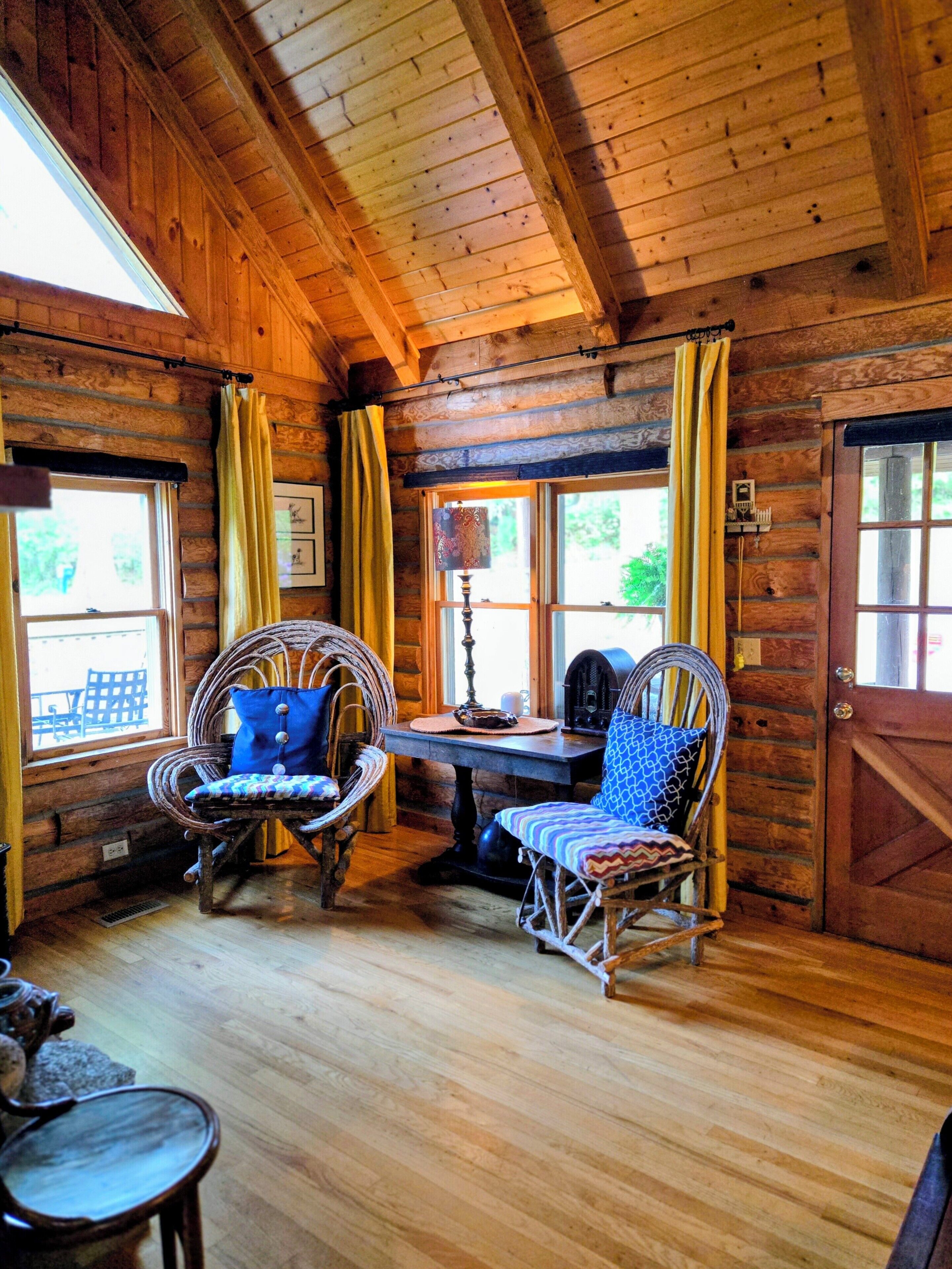 A log cabin interior with wooden walls, a dining table, wicker chairs, and a large window.