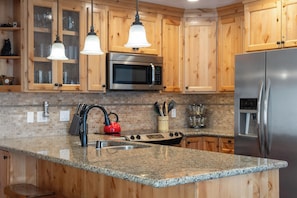A kitchen with wooden cabinets, a stainless steel refrigerator, a microwave, a granite countertop, and pendant lights. - Quiet Walk to Slopes Condo 660 (Mammoth Lakes)