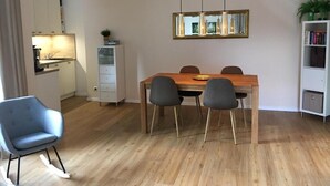 A dining area with a wooden table, four chairs, a hanging light fixture, a bookshelf, and a kitchen in the background. - Vacation apartment Gute Zeit Hahnenklee (Goslar)
