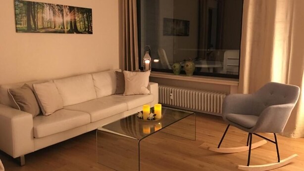 A modern living room with a sofa, a rocking chair, a glass coffee table with candles, and a large window with curtains. - Vacation apartment Gute Zeit Hahnenklee (Goslar)