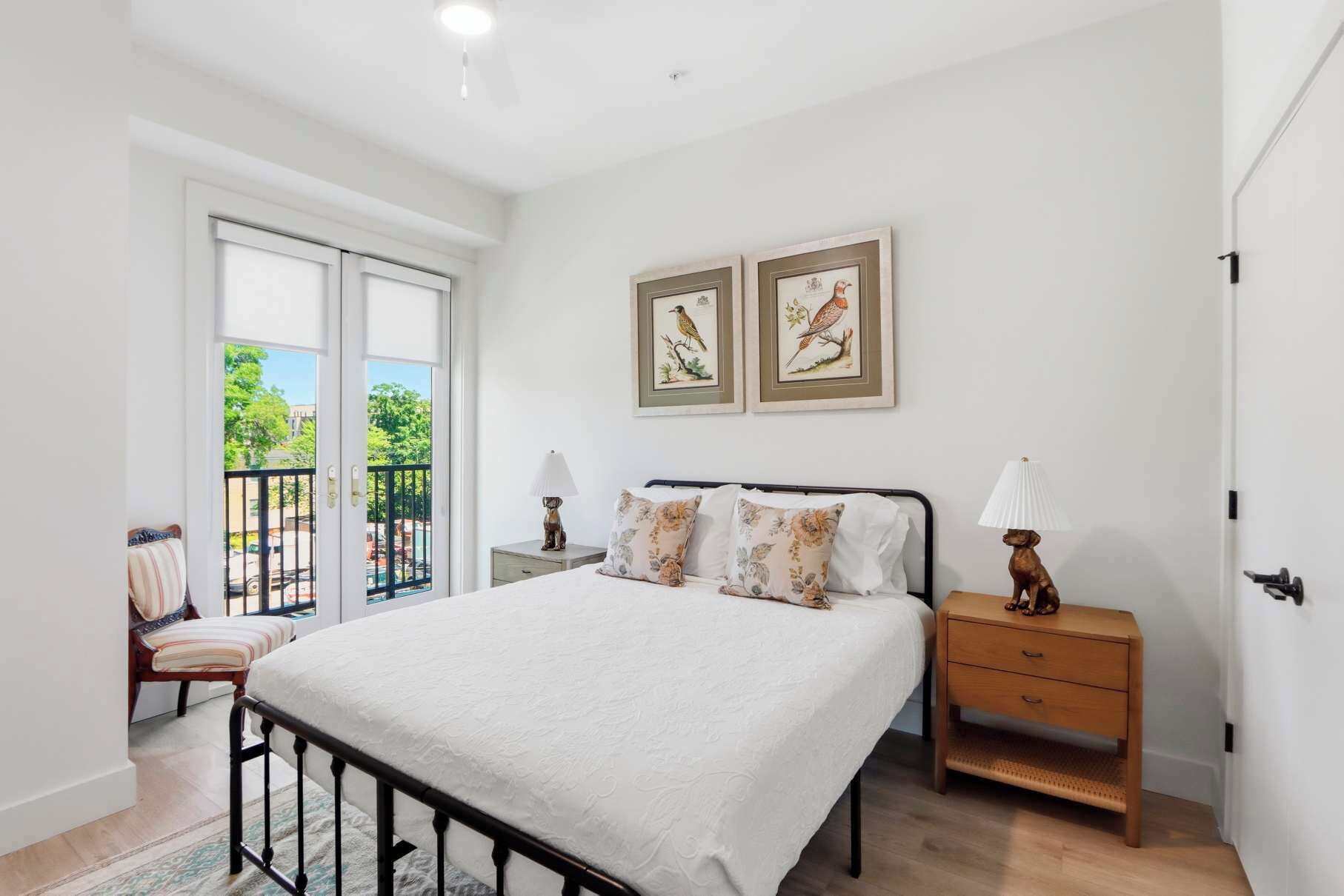 A bedroom with a bed, a nightstand, a chair, a balcony, and two framed pictures on the wall.