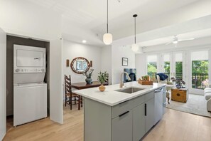A modern kitchen with a central island, a dining area with a chair and table, and a view of the outdoors through large windows. - The Jardin Wisteria Suite | Near Downtown (Savannah)