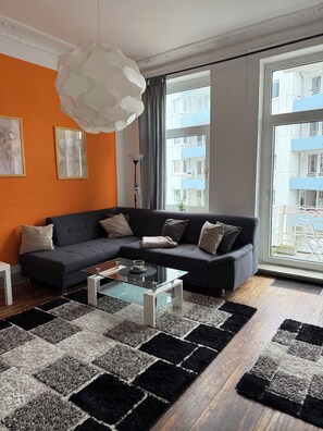 A modern living room with a sectional sofa, a glass coffee table, and a view of a building through large windows. - 4-room apartment in Cuxhaven on the dyke (Cuxhaven)