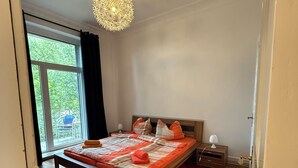 A bedroom with a bed, a nightstand, a lamp, a red rug, and a view through a window. - 4-room apartment in Cuxhaven on the dyke (Cuxhaven)
