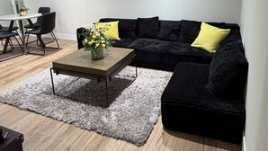 Ein modernes Wohnzimmer mit einem schwarzen Sofa, einem Couchtisch mit Blumen in einer Vase, einem grauen Teppich und einem Essbereich mit Stühlen und einem Tisch.