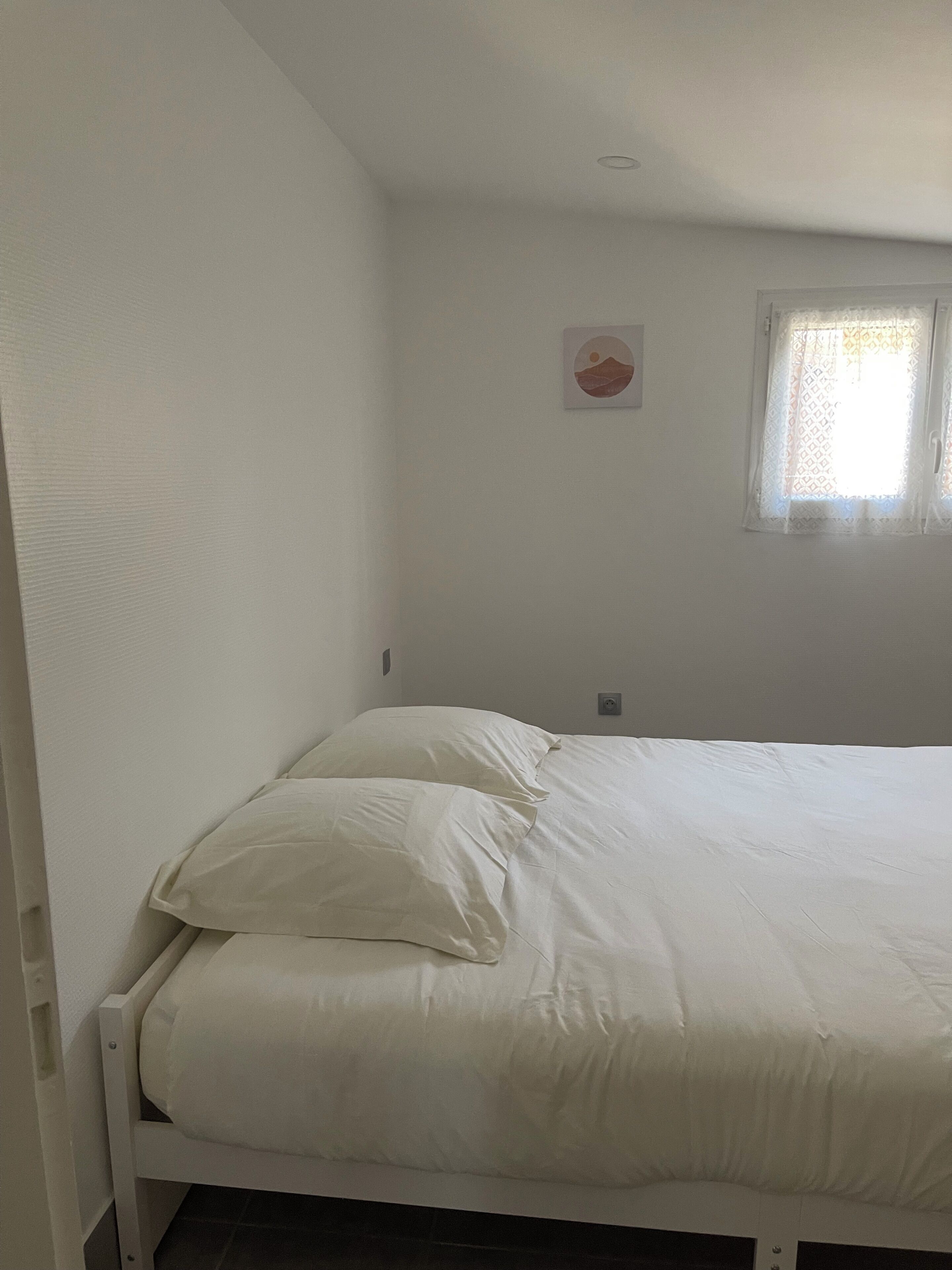 A single bed with white bedding in a room with white walls, a small window with patterned curtains, and a wall-mounted light fixture.