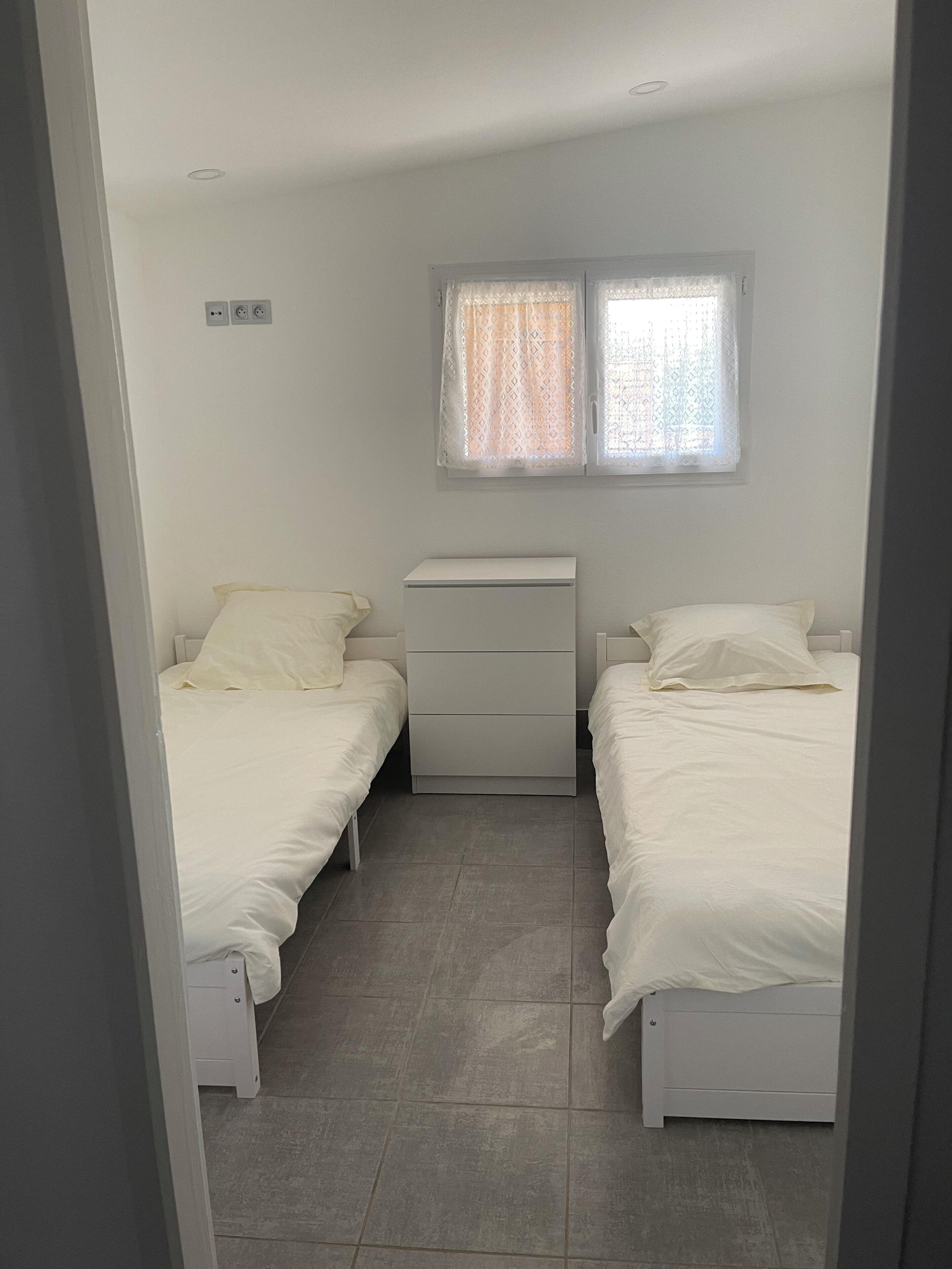 Une petite chambre propre, avec deux lits, une table de chevet et une fenêtre agrémentée de rideaux à motifs.