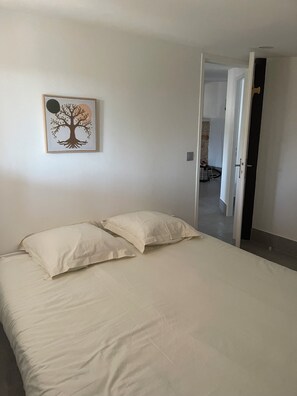 Una habitación con cama, almohadas, un cuadro en la pared y una puerta abierta que da a otra habitación.