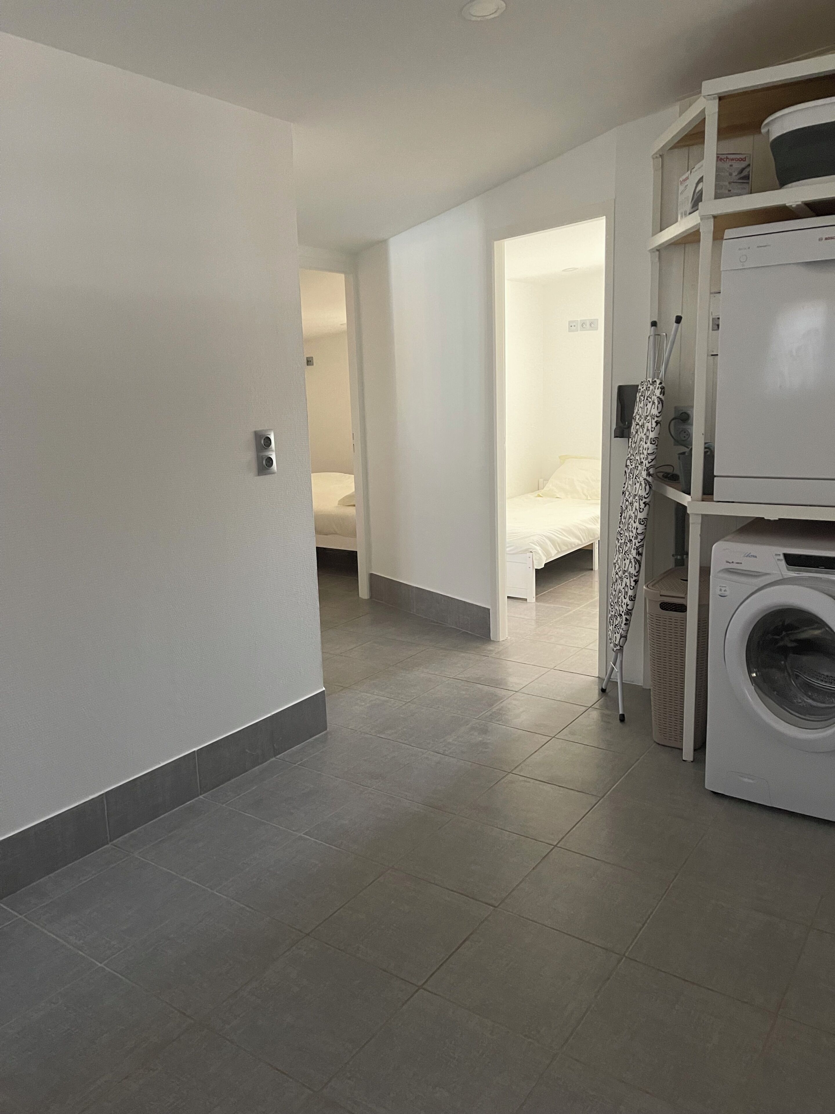 A laundry area with a washing machine, a drying rack, and a bed in the background.