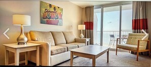 A hotel room with a sofa, two side tables, a coffee table, a chair, and a large window with curtains. - Cozy 1-bedroom condo limited Ocean view balcony in adorable North Myrtle Beach (North Myrtle Beach)