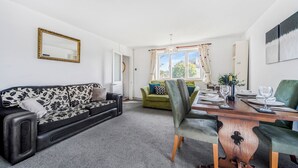 En stue med en sofa i sort og hvidt mønster, en grøn sofa, et træspisebord med stole og et stort vindue med gardiner.