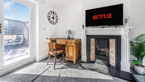 En stue med pejs, et skrivebord med stol, et fjernsyn der viser 'NETFLIX', et ur og udsigt gennem en glasdør.