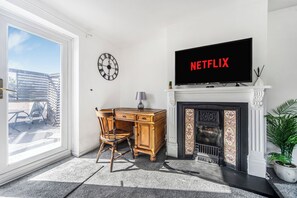 Een woonkamer met een open haard, een bureau met stoel, een tv waarop 'NETFLIX' staat, een klok en uitzicht door een glazen deur.