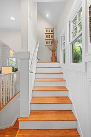 A staircase with wooden steps and white walls, a framed photo collection on the wall, and a window with a view of greenery. - Overlooking the 13th tee of the Bald Head Island Club golf course (Bald Head Island)