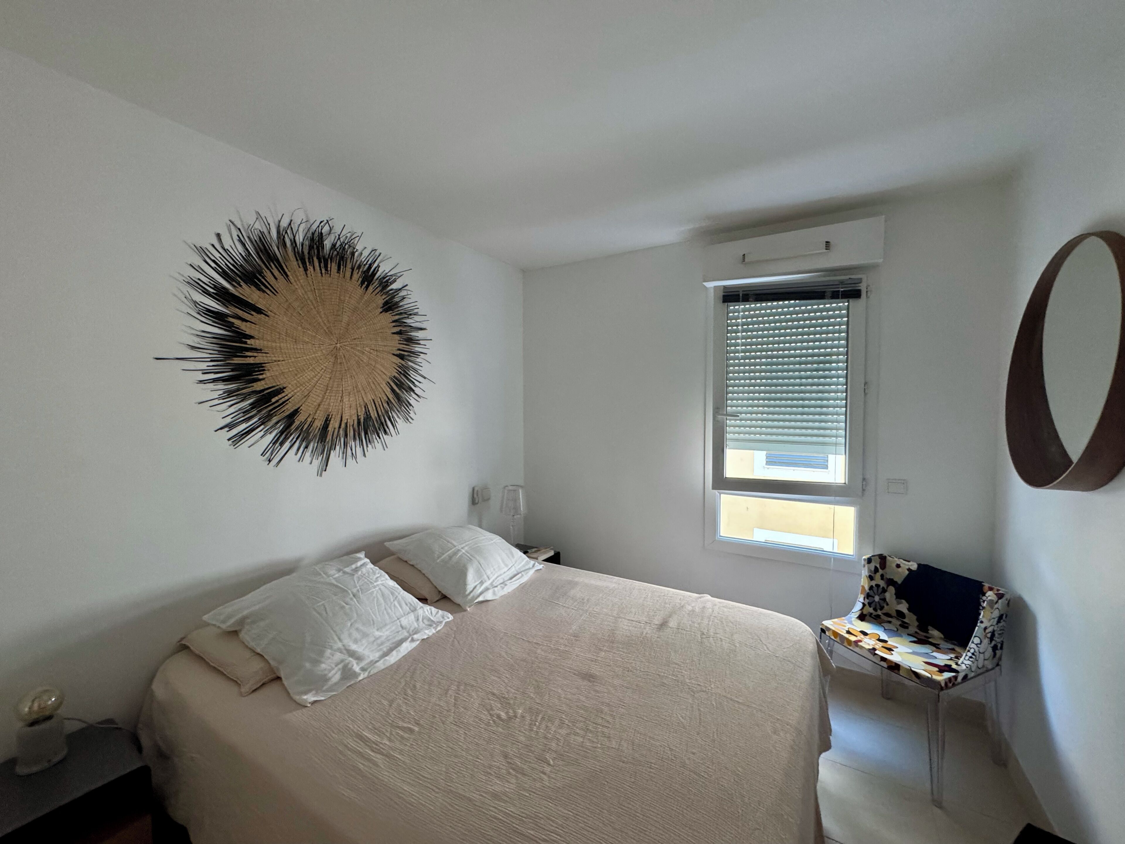 Une chambre à coucher avec un lit, un élément de décoration murale rond, une fenêtre avec des stores, une chaise et un miroir.