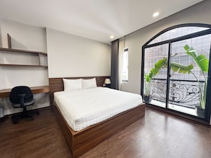 Une chambre d’hôtel avec un lit en bois, un bureau, une chaise, une fenêtre donnant sur un balcon et une plante en pot.