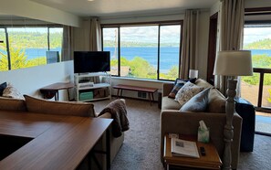 Ein Wohnzimmer mit Fernseher, Sofa und Tisch, große Fenster mit Blick auf einen See und Gebäude.